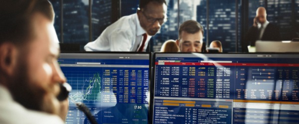 A stock trader at his desk, keeping an eye on the market and talking to an investor.